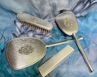 May include: Three silver-toned hair care tools on a blue and white patterned fabric. The tools include a brush with natural bristles, a comb, and two oval-shaped hand mirrors.
