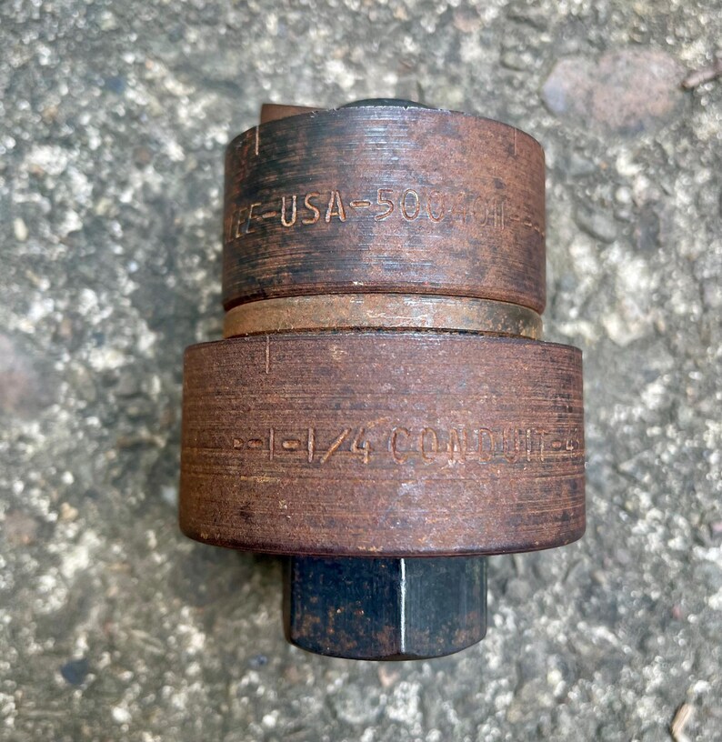 May include: A close-up of a rusty metal tool with the text "E-USA-500" and "1-1/4 CONDUIT" etched into the metal. The tool has a cylindrical shape with a hexagonal base.