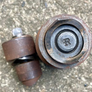 May include: Close-up of two rusty metal tools. One tool is cylindrical with a hexagonal opening, and the other is a smaller, multi-part tool with a bolt. The larger tool has the letter "R" stamped on its center.