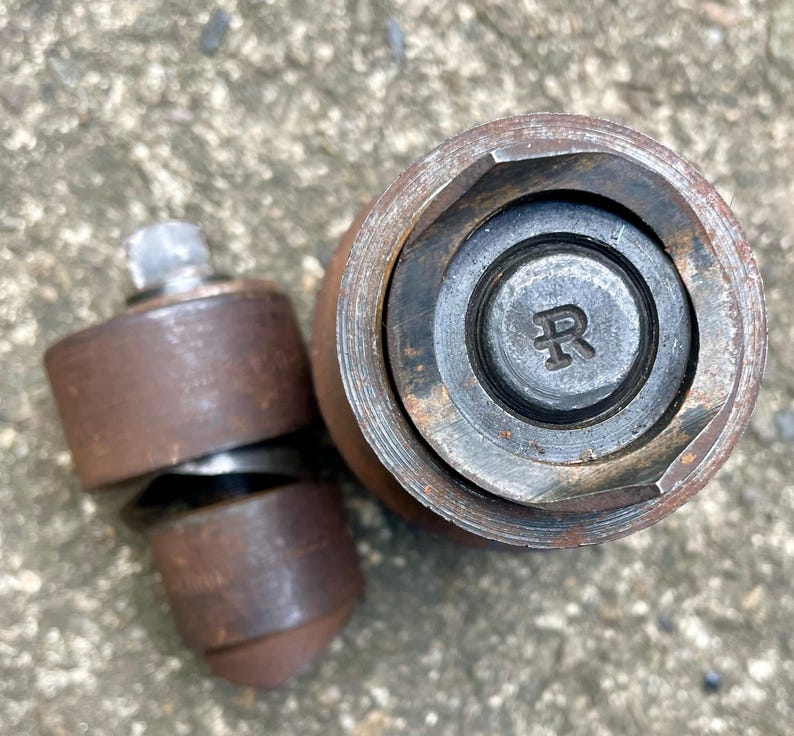 May include: Close-up of two rusty metal tools. One tool is cylindrical with a hexagonal opening, and the other is a smaller, multi-part tool with a bolt. The larger tool has the letter "R" stamped on its center.