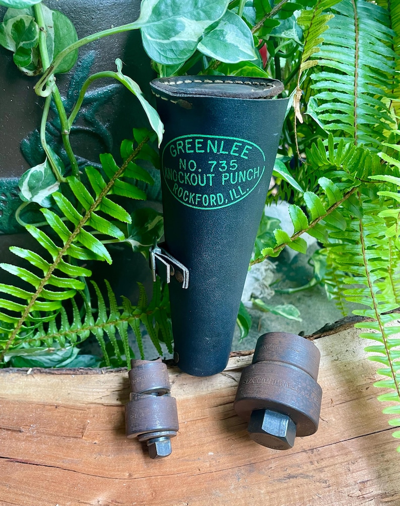 May include: A black leather Greenlee No. 735 Knockout Punch tool set with a holder. The holder has the Greenlee logo in green. Two metal punch components are next to the holder. The tools are on a wooden surface, surrounded by green plants.