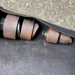 May include: A black leather tool roll with brown and metal tools inside. The tools include various sizes of metal rings and a large bolt. The roll is open, displaying the tools, and has white stitching.