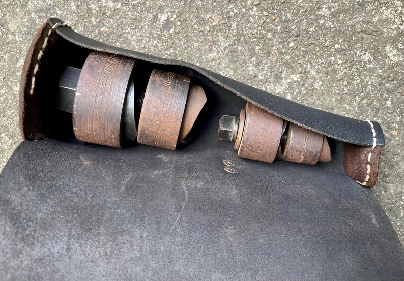 May include: A black leather tool roll with brown and metal tools inside. The tools include various sizes of metal rings and a large bolt. The roll is open, displaying the tools, and has white stitching.