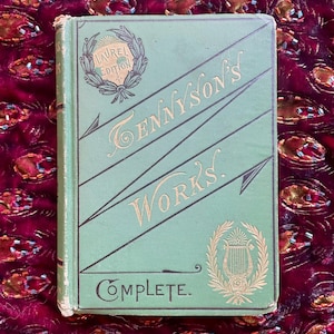 May include: A vintage book with a teal cover featuring gold lettering that reads "Tennyson's Works. Complete." The cover includes laurel wreath and harp illustrations. The book's edges show signs of wear.