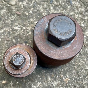 May include: Two weathered metal tools with hexagonal and square bolt heads. The larger tool has the text "GREENLEE" and numbers on the bolt head. The tools are circular and cylindrical, showing signs of rust and wear.