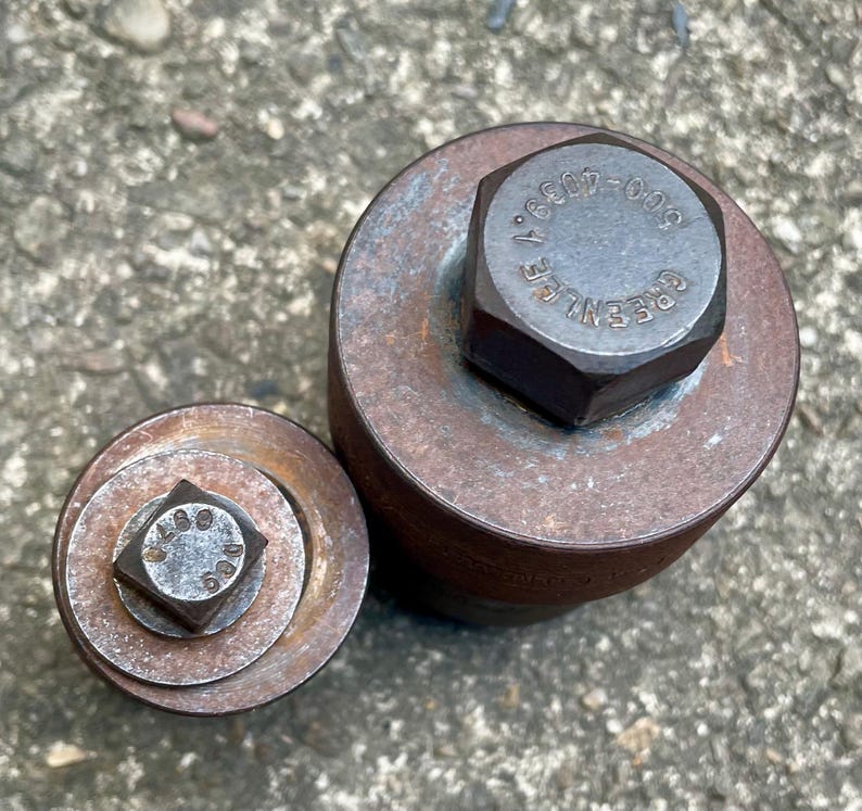 May include: Two weathered metal tools with hexagonal and square bolt heads. The larger tool has the text "GREENLEE" and numbers on the bolt head. The tools are circular and cylindrical, showing signs of rust and wear.