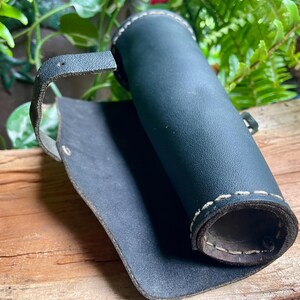 May include: A rolled-up, dark blue leather quiver with white stitching and a strap. The quiver is open, revealing the interior. The background includes greenery and a wooden surface.