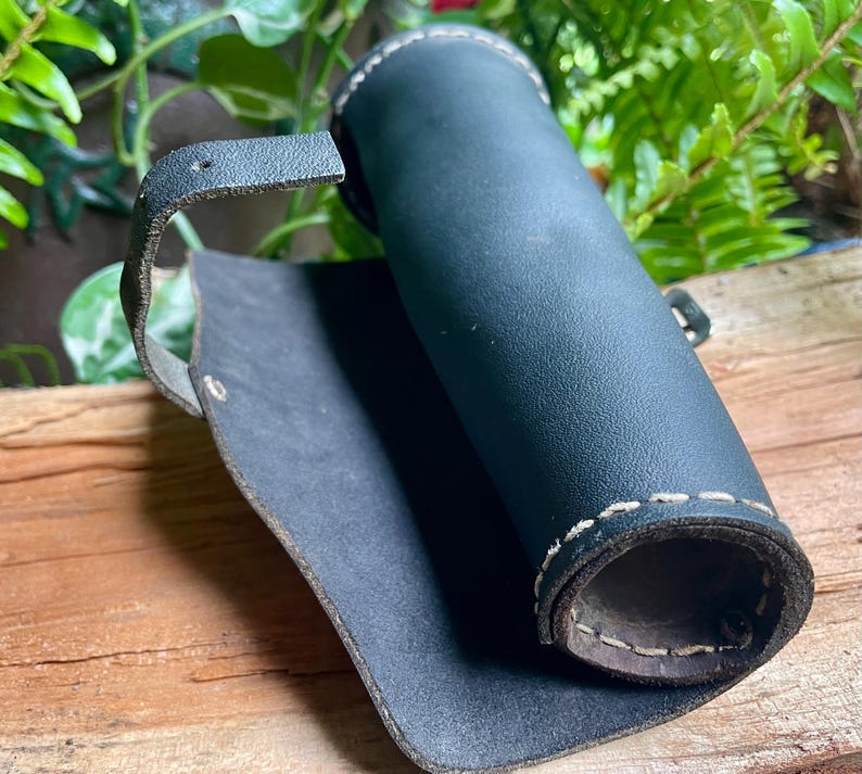 May include: A rolled-up, dark blue leather quiver with white stitching and a strap. The quiver is open, revealing the interior. The background includes greenery and a wooden surface.