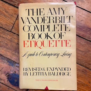 May include: A vintage copy of "The Amy Vanderbilt Complete Book of Etiquette" with a cream-colored cover. The title is in black, with "Etiquette" in red. The book is a guide to contemporary living, revised and expanded by Letitia Baldrige.