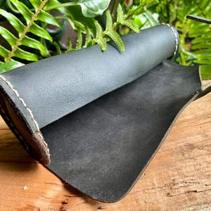 May include: A black leather pen holder with a rolled design and a curved handle. The edges are stitched with a light-colored thread, and the interior is a darker shade of black. The pen holder is displayed on a wooden surface.