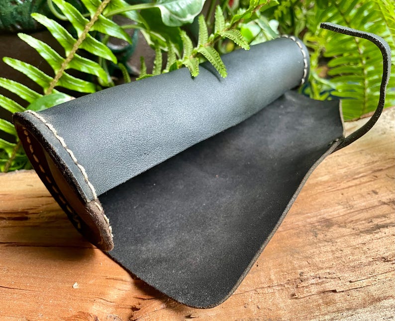 May include: A black leather pen holder with a rolled design and a curved handle. The edges are stitched with a light-colored thread, and the interior is a darker shade of black. The pen holder is displayed on a wooden surface.