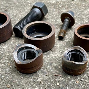 May include: A collection of metal hardware components, including bolts, nuts, and cylindrical pieces. The items are various sizes and show signs of wear, with a mix of dark and rusty brown tones. The components are arranged on a rough, gray surface.