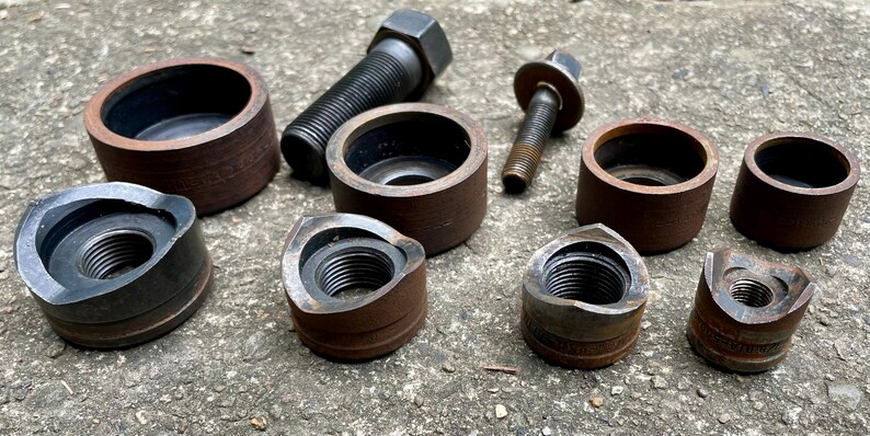 May include: A collection of metal hardware components, including bolts, nuts, and cylindrical pieces. The items are various sizes and show signs of wear, with a mix of dark and rusty brown tones. The components are arranged on a rough, gray surface.