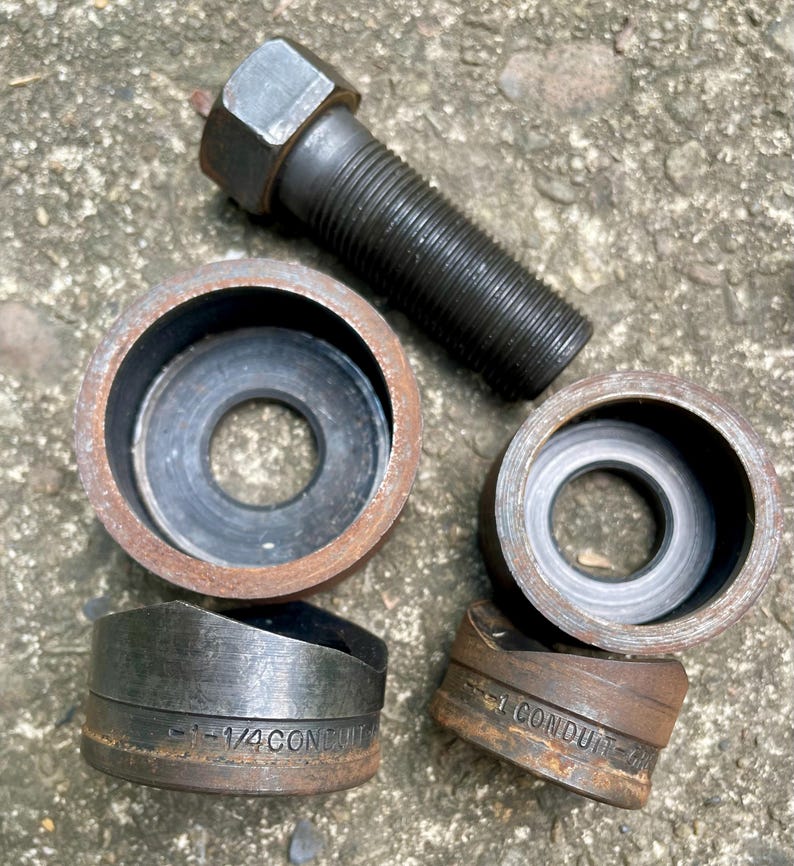 May include: A collection of metal tools, including a bolt and several conduit punches. The punches are labeled with size specifications. The tools have a weathered, used appearance, suggesting they are for industrial or construction use.