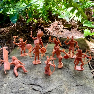 May include: A collection of small, orange-brown plastic toy Native American figures. The figures are in various poses, some holding weapons like bows and arrows, tomahawks, and shields. The toys are arranged on a gray stone surface, with green foliage in the background.