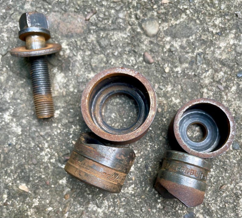 May include: A collection of rusty metal tools, including a bolt with a hexagonal head and several cylindrical pieces. The pieces have a dark, aged appearance, suggesting they are well-used. Some pieces have text that is difficult to read.