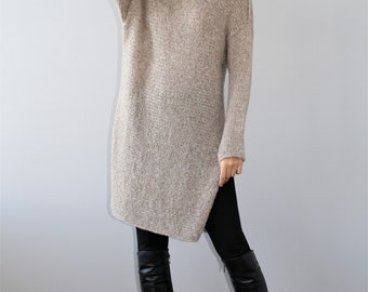 May include: A long, beige knitted sweater dress with a loose fit and a slightly asymmetrical hem. The model is wearing black leggings and tall black boots.