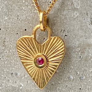 Könnte beinhalten: Goldene herzförmige Halskette mit einem roten Edelstein in der Mitte. Der Anhänger hat ein Sonnenstrahlendesign und ist an einer goldenen Kette befestigt.