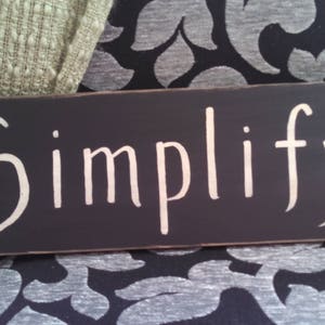 May include: Black wooden sign with white lettering that reads "Simplify".