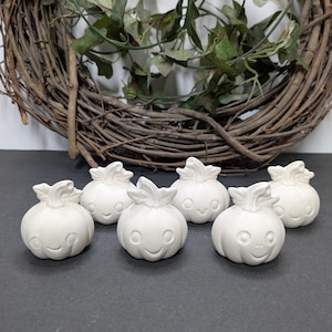 May include: A collection of six small, white ceramic figurines shaped like smiling pumpkins. Each pumpkin has a carved face and a leafy top. They are arranged on a dark surface, with a woven wreath and greenery in the background.