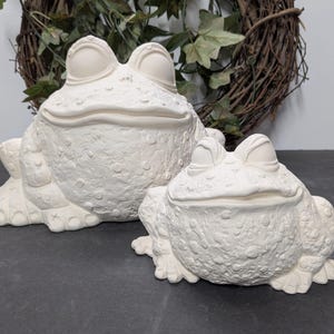 May include: Two unpainted ceramic frog figurines. The larger frog is in the background, and the smaller frog is in the foreground. Both frogs have a textured surface and are in a sitting position. A decorative wreath is in the background.