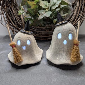 May include: Two ceramic ghost figurines with a crackled white finish, each wearing a small black witch hat and holding a miniature straw broom. The ghosts feature illuminated blue eyes and mouths, creating a glowing effect for seasonal decor.