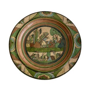 May include: A round ceramic plate featuring a hand-painted scene of a person carrying a bundle of wood near cacti and trees. The rim is decorated with concentric rings and repeating wave-like patterns in shades of green, brown, and tan.