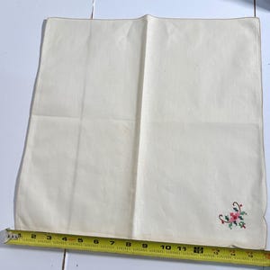 May include: Off-white linen handkerchief with a small embroidered floral design in the corner. The design includes red, pink, and green elements. The handkerchief measures approximately 14 inches long.