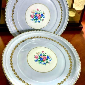 Può includere: Una pila di piatti in ceramica azzurra con un motivo floreale. Ogni piatto presenta un cerchio centrale color crema con un bouquet di fiori rosa, blu e bianchi. I bordi sono decorati con motivi a foglia d'oro e un bordo perlato.