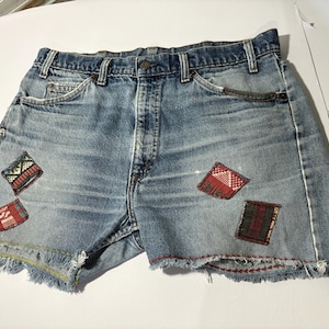Peut inclure: Un short en jean bleu avec des bords effilochés et diverses patchs en tissu coloré cousues dessus. Les patchs sont de formes et de tailles différentes, et certaines ont des motifs géométriques.