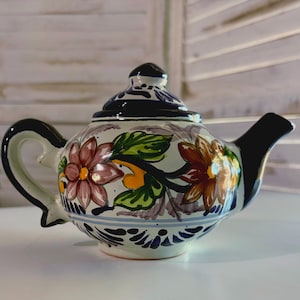 May include: A small white ceramic teapot with a black handle and spout. The teapot is decorated with hand-painted floral designs in shades of pink, orange, yellow, and green. The teapot has a blue and white trim around the top and bottom.