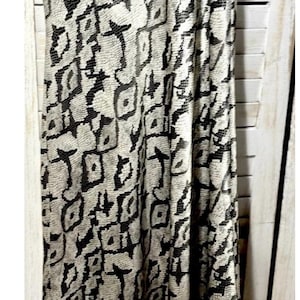 May include: A floor-length dress with a black and beige snakeskin pattern. The dress has thin straps and a flowing silhouette. The fabric drapes down, creating a vertical visual effect. The dress is displayed against a white shuttered background.