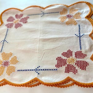 Puede incluir: Mantel de mesa blanco de ganchillo con borde festoneado y diseños florales bordados en tonos rojos, amarillos y azules.
