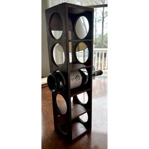Pode incluir: Um suporte para vinhos retangular e alto, feito de madeira castanho-escura, com cinco aberturas circulares para armazenamento de garrafas. Uma garrafa de vinho de vidro com um rótulo branco está posicionada horizontalmente no compartimento central.