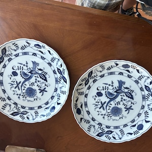May include: Two white ceramic plates with a blue floral and bird design. The plates have a scalloped edge and a detailed pattern featuring a bird, flowers, and foliage. The plates are on a wooden surface.