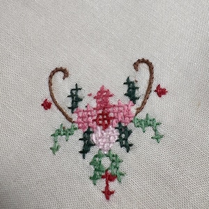 May include: A close-up of a cross-stitch floral design. The design features a central flower in shades of pink and red, surrounded by green leaves and accents. The embroidery is set against a light beige fabric.