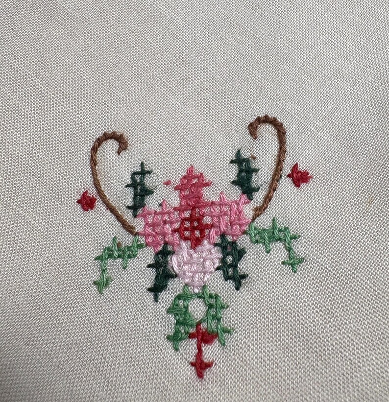 May include: A close-up of a cross-stitch floral design. The design features a central flower in shades of pink and red, surrounded by green leaves and accents. The embroidery is set against a light beige fabric.