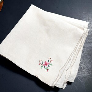 May include: A stack of off-white linen napkins with a scalloped edge. Each napkin features a small, embroidered floral design in pink, green, and red. The napkins are neatly folded and stacked, showcasing the delicate embroidery.