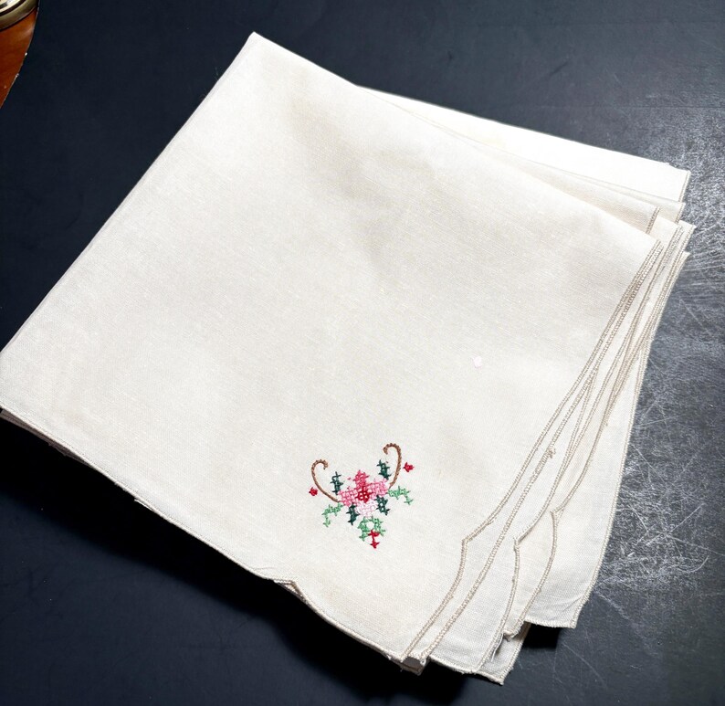 May include: A stack of off-white linen napkins with a scalloped edge. Each napkin features a small, embroidered floral design in pink, green, and red. The napkins are neatly folded and stacked, showcasing the delicate embroidery.