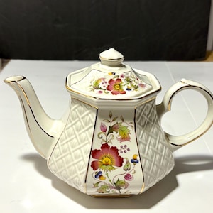 May include: White teapot with a floral design and a gold trim. The teapot has a woven pattern and a lid with a floral design.