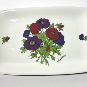 May include: Rectangular white ceramic dish with a floral design. The design features a bouquet of red, purple, and blue flowers with green leaves. The dish has rounded corners and a signature in the bottom right corner.
