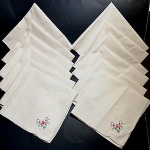 May include: A set of cream-colored cloth napkins, each with a scalloped edge and embroidered floral design in red, pink, and green. The napkins are neatly arranged on a dark surface, showcasing their delicate details.