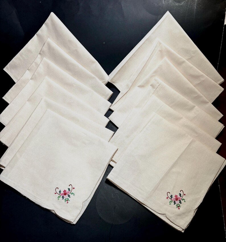 May include: A set of cream-colored cloth napkins, each with a scalloped edge and embroidered floral design in red, pink, and green. The napkins are neatly arranged on a dark surface, showcasing their delicate details.