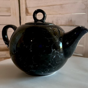 Puede incluir: Tetera negra con un patrón de mármol. La tetera tiene un cuerpo redondo y una pequeña asa.