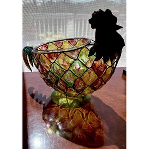 May include: A decorative glass bowl with a pineapple design. The bowl features a grid pattern with red, yellow, and green glass pieces. It has a black metal rooster silhouette on the side and a green handle. The bowl sits on a round, green base.