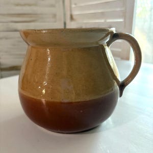 May include: A brown and tan ceramic pitcher with a rounded body and a handle. The pitcher has a glazed finish.