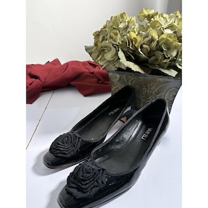 Op de afbeelding: Een paar zwarte lakleren ballerina's met een rozenversiering op de neus. De schoenen zijn van het merk "VAN ELI". Een rood kledingstuk en een decoratieve plant staan op de achtergrond.