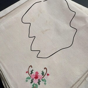 May include: A beige linen napkin with a floral embroidery design. The embroidery features a pink flower with green and red accents, and brown curved stems. The napkin has a scalloped edge.