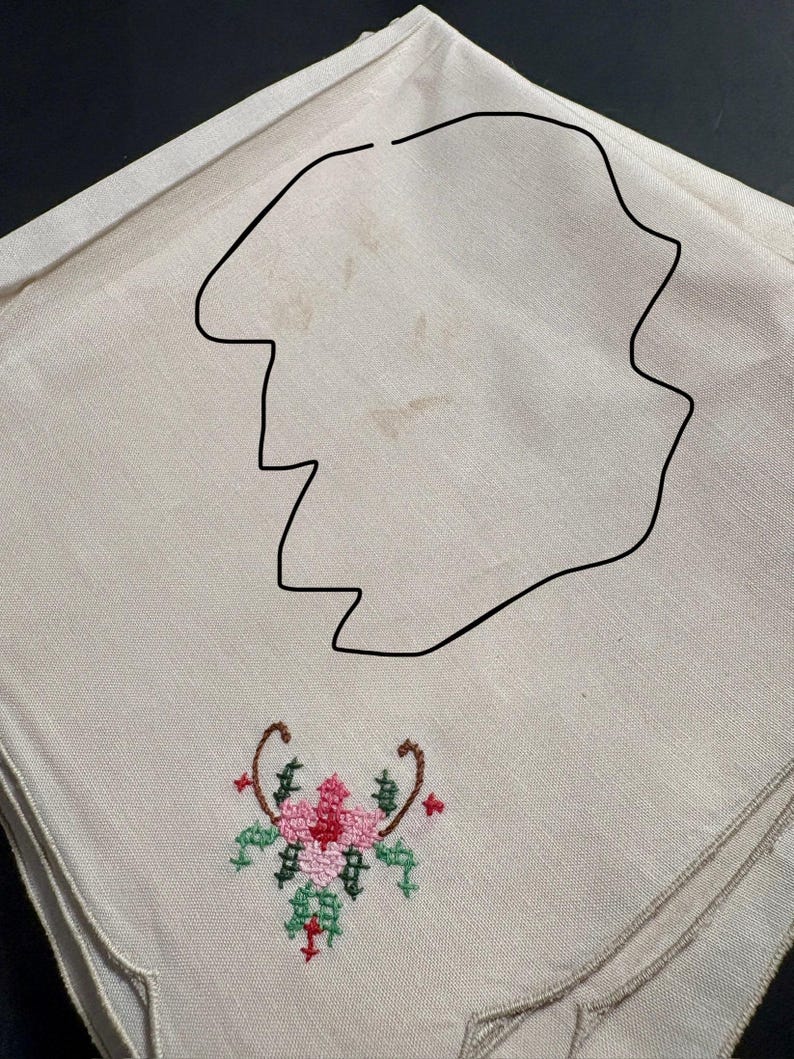 May include: A beige linen napkin with a floral embroidery design. The embroidery features a pink flower with green and red accents, and brown curved stems. The napkin has a scalloped edge.