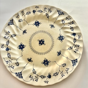 May include: A white ceramic dinner plate with a blue floral pattern. The plate has a scalloped edge and a raised center.